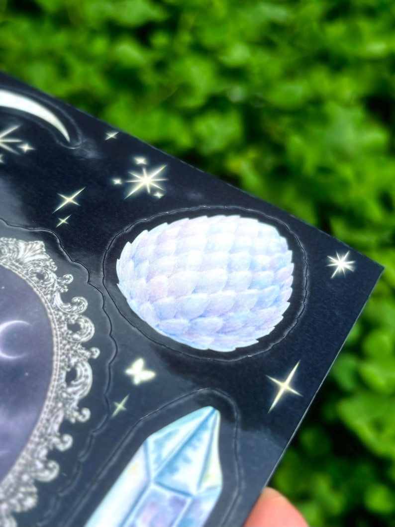 May include: A sheet of stickers with a celestial theme. The stickers feature a moon, stars, a crystal, and a round, feathery design in shades of blue and white. The background is a dark blue with a glossy finish.