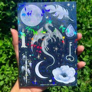Może przedstawiać: Holograficzna arkusz naklejek z ilustracjami fantasy. Projekty obejmują smoka, księżyc, pi&oacute;ro, klucz, miecz, kwiat i klejnot, w odcieniach bieli i srebra na ciemnoniebieskim tle. Widoczny jest tekst "PEACH CAT PRINTS".