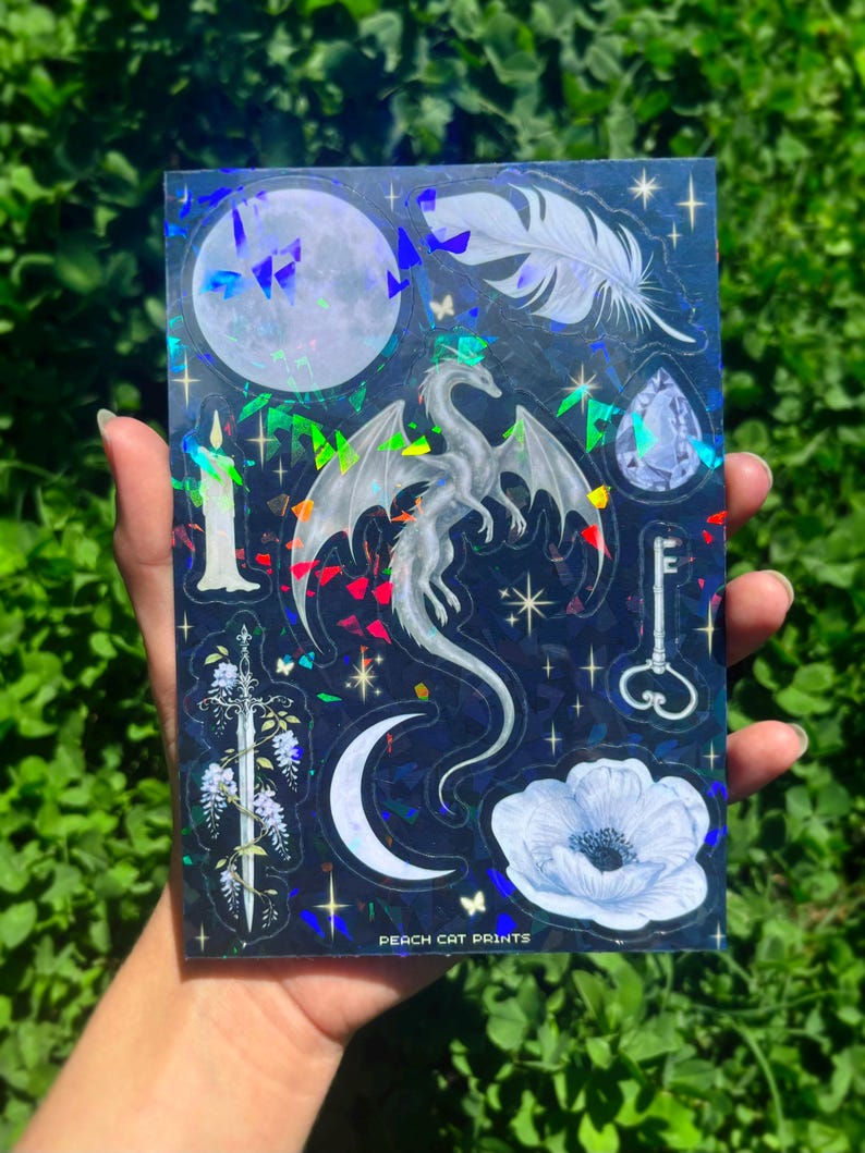 Może przedstawiać: Holograficzna arkusz naklejek z ilustracjami fantasy. Projekty obejmują smoka, księżyc, pi&oacute;ro, klucz, miecz, kwiat i klejnot, w odcieniach bieli i srebra na ciemnoniebieskim tle. Widoczny jest tekst "PEACH CAT PRINTS".