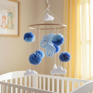 May include: A baby mobile featuring a light blue elephant, fluffy blue pom-poms, and white cloud shapes. The mobile hangs from a wooden ring and is suspended above a white crib. The nursery decor has a soft, calming aesthetic.