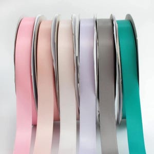 May include: A selection of satin ribbon spools in pastel shades: pink, peach, cream, lavender, grey, and teal. The ribbons are neatly rolled, with the edges of the ribbon visible. Suitable for crafts and gift wrapping. Each spool is approximately 2.5 cm wide.