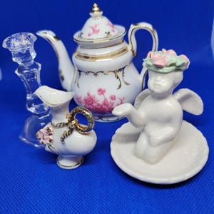 Puede incluir: Un juego de té de porcelana blanca con un diseño floral. El juego incluye una tetera, una jarra de crema, un azucarero y una pequeña figura de ángel con una corona de flores.