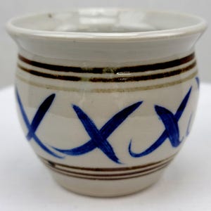 May include: A small, off-white ceramic pot with a rounded shape. The pot features three dark brown horizontal stripes near the top and bottom, and blue brushstroke 'X' designs. The rim is a lighter off-white color.