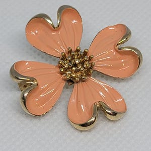 Puede incluir: Broche en forma de flor con cuatro pétalos color melocotón y un borde dorado. El centro del broche tiene un grupo de pequeñas cuentas doradas. El broche es probablemente de metal y esmalte.
