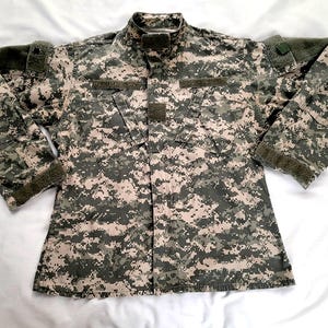 Pode incluir: Uma jaqueta militar de camuflagem com fecho de botões e mangas compridas. A jaqueta tem um remendo na manga direita e um bolso no peito esquerdo.