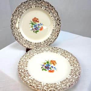 Puede incluir: Dos platos de cerámica blanca con ribetes dorados y diseños florales. Los platos tienen un borde festoneado y un patrón floral en el centro. Las flores son de color naranja, azul y morado.