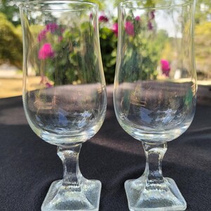 May include: Two clear glass goblets with square bases. The goblets have a rounded, flared bowl and a short stem.