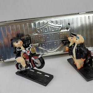 Puede incluir: Figuras de cerdos vestidos con atuendos de moteros, montando motocicletas, con un logotipo de Harley-Davidson en el fondo. Las motocicletas son rojas y negras, y las figuras están sobre una base negra.