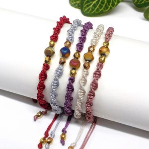 Può includere: Cinque braccialetti intrecciati con perline di vetro iridescenti e dettagli dorati. I braccialetti sono in tonalità di rosso, argento, viola, crema e rosa.