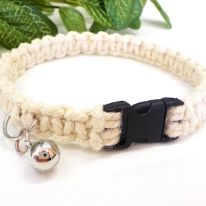 Könnte beinhalten: Ein weißes Katzenhalsband aus Baumwollseil mit einer schwarzen Kunststoffschnalle und einer silbernen Glocke.