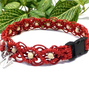 Könnte beinhalten: Rotes Makramee-Halsband für Hunde mit Holzperlen und einer schwarzen Kunststoffschnalle. Ein silberner Anhänger mit dem Buchstaben "M" hängt daran. Das Halsband ist leuchtend rot.
