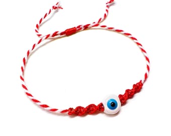 Könnte beinhalten: Ein rot-weiß geflochtenes Armband mit einem weißen und blauen Augenanhänger. Das Armband besteht aus gedrehten roten und weißen Fäden, mit einem zentralen Abschnitt aus geknotetem rotem Faden. Der Augenanhänger hat eine blaue Iris.