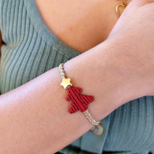 Pode incluir: Um bracelete de árvore de Natal vermelho e dourado. O bracelete é feito de um cordão tecido e apresenta um amuleto de árvore de Natal vermelho com uma estrela dourada no topo.