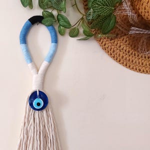 Pode incluir: Um pendurado de parede de franja branca com um amuleto de olho azul e preto. A franja é feita de fio de algodão e tem um amuleto de olho azul e preto na parte inferior. O amuleto de olho é um símbolo tradicional de proteção e boa sorte.