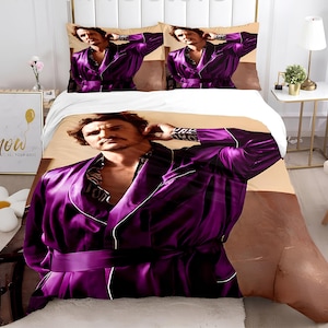 May include: A purple satin robe with a white trim and a belt. The robe is worn by a man with a beard and mustache. The background is a beige and brown duvet cover.