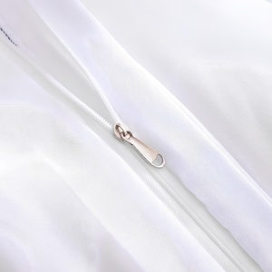 Puede incluir: Primer plano de una cremallera blanca con un tirador de cremallera plateado. La cremallera est&aacute; cosida en una tela blanca.