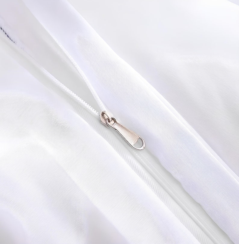 Puede incluir: Primer plano de una cremallera blanca con un tirador de cremallera plateado. La cremallera est&aacute; cosida en una tela blanca.