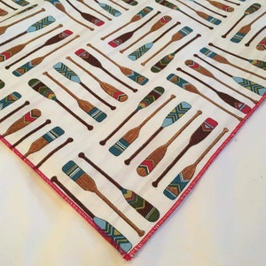 Peut inclure: Tissu blanc avec un motif répété de pagaies de canoë marron, turquoise, rouge et vert. Les pagaies présentent des motifs géométriques. Le tissu a une bordure cousue rouge.