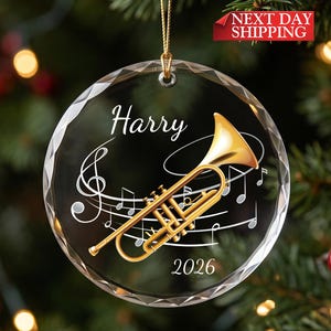 May include: A circular, faceted glass ornament featuring a gold-colored trumpet illustration, white musical notes, and the text Harry and 2026. The ornament hangs from a gold string against a blurred background of green pine needles and lights.