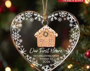 May include: A heart-shaped clear acrylic ornament featuring a white printed snowflake border, a central gingerbread house illustration, and the text "Our First Home 581 S WILLOWBROOK LN PHOENIX AZ 2026" suspended by a thin gold cord.