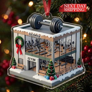May include: A two-story gym building Christmas ornament featuring a black dumbbell on the roof, a wreath on the side, and a decorated Christmas tree by the entrance. The building has glass walls, interior gym equipment, and the text GEORGE GYM.