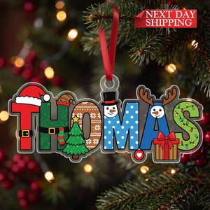 May include: A personalized Christmas ornament featuring the name THOMAS in large, colorful letters. Each letter incorporates holiday motifs including a Santa hat, elf hat, Christmas tree, snowman, reindeer antlers, and string lights.