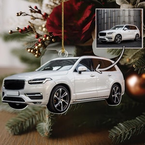 May include: White SUV car ornament hanging from a gold string. The ornament is a detailed replica of a modern SUV with black accents and a black grille. The ornament is hanging from a Christmas tree.