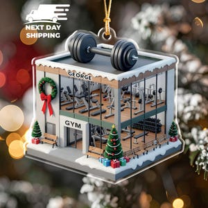 May include: A clear acrylic Christmas ornament shaped like a gym. The ornament features a barbell on top, a wreath, and a Christmas tree. The word "GEORGE" is above the gym, and "GYM" is on the front. The gym has exercise equipment inside.