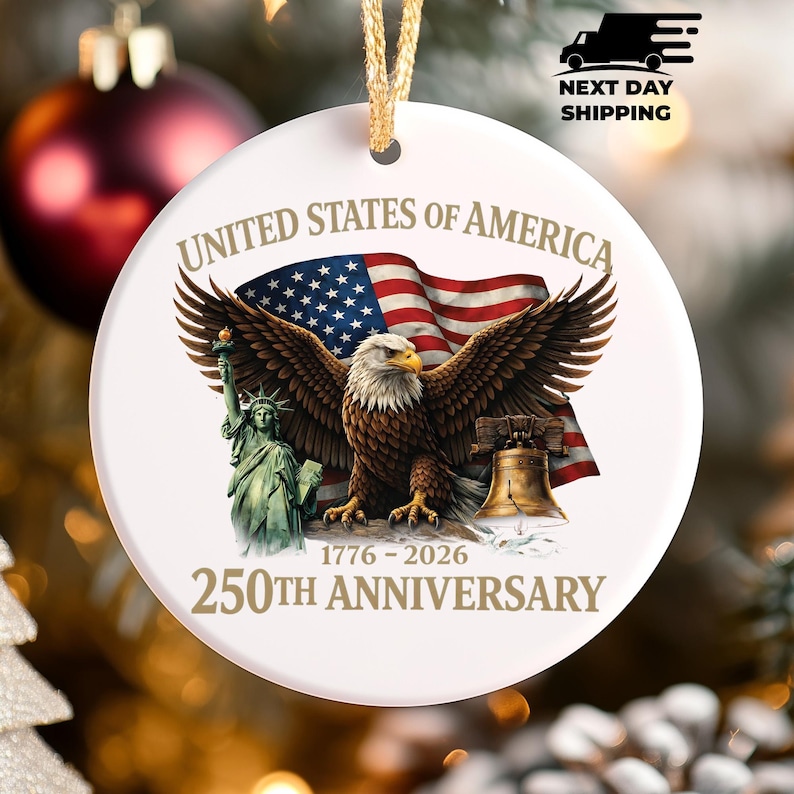 Puede incluir: Adorno redondo de cer&aacute;mica blanca con una cuerda dorada para colgar. Presenta un &aacute;guila calva, la bandera estadounidense, la Estatua de la Libertad y la Campana de la Libertad. El texto incluye "UNITED STATES OF AMERICA" y "250TH ANNIVERSARY 1776-2026."