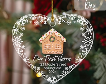 May include: Heart-shaped clear glass ornament with a gingerbread house design. The ornament has a snowflake border and the text "Our First Home" with an address and the year 2024.