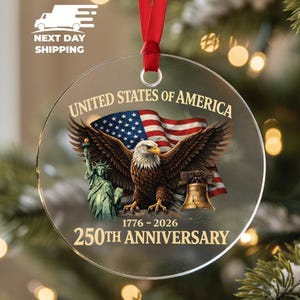 Puede incluir: Adorno navide&ntilde;o redondo y transparente con una cinta roja. El adorno presenta una bandera estadounidense, un &aacute;guila con las alas extendidas, la Estatua de la Libertad y la Campana de la Libertad. El texto dice "United States of America" y "250th Anniversary 1776-2026."