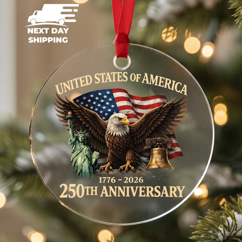 Puede incluir: Adorno navide&ntilde;o redondo y transparente con una cinta roja. El adorno presenta una bandera estadounidense, un &aacute;guila con las alas extendidas, la Estatua de la Libertad y la Campana de la Libertad. El texto dice "United States of America" y "250th Anniversary 1776-2026."