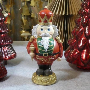 Könnte beinhalten: Eine dekorative Weihnachts-Nussknacker-Figur in roter und goldener Uniform, die ein Zepter hält. Die Figur hat einen weißen Bart und eine rote Krone. Zwei rote Weihnachtsbaumschmuck aus Glas befinden sich auf beiden Seiten.