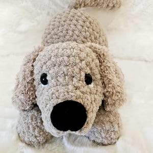 May include: A crocheted plush dog toy in a light brown color with black eyes and a black nose. The dog is lying on a white fluffy surface.