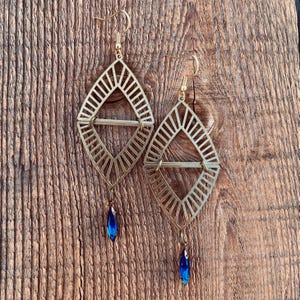 May include: A pair of gold-colored earrings with a geometric diamond-shaped design. Each earring features a blue teardrop-shaped crystal pendant. The earrings are displayed on a wooden surface.
