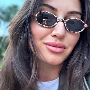 May include: Oval sunglasses featuring a dark, multi-coloured floral pattern on the frames and dark tinted lenses. The frames have a rounded shape with a bridge connecting the two lenses, providing a distinct look with a colourful, patterned finish.