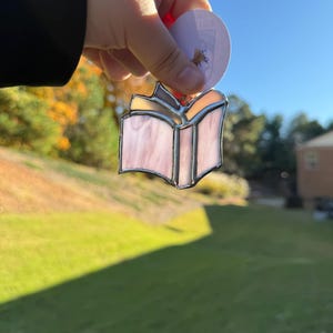 May include: A stained glass ornament shaped like an open book with pale pink, translucent glass panels held together by silver-colored metal solder lines. The ornament is suspended by a small piece of twine and features a circular white tag.