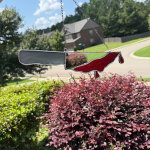 May include: A stained glass suncatcher shaped like a cleaver knife with a grey blade and a red stained glass section on the edge that resembles dripping blood. The piece is held by a thin metal chain and features dark metal solder lines.