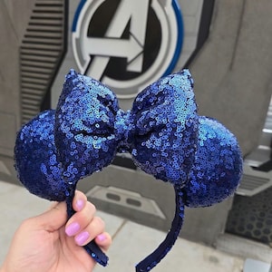 Puede incluir: Diadema de orejas de Minnie Mouse de lentejuelas azules con un gran lazo. La diadema está delante de una pared gris con un logotipo de los Vengadores azul y blanco.