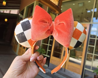 May include: Orange and black checkered mouse ears with a large orange bow. The headband is orange with white stripes. The ears have a black and white checkered pattern and an orange and white checkered pattern. The bow is made of a soft, velvet-like material.