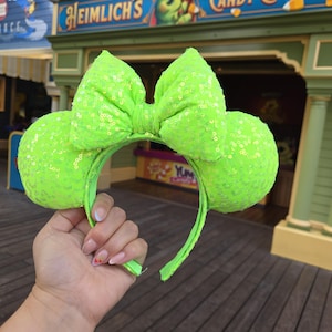 Puede incluir: Diadema de orejas de Minnie Mouse verde lima con lentejuelas y un lazo grande. La diadema se sostiene frente a una tienda con el letrero "Heimlich's Candy Corn Toss". Las orejas y el lazo están cubiertos de lentejuelas.