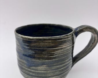 Op de afbeelding: Handgemaakte keramische mok met een donkerblauwe en olijfgroene glazuur. De mok heeft een gestructureerd, horizontaal gestreept patroon en een gebogen handvat. De binnenkant is diepblauw en de basis heeft een lichtroze tint. Voor koffie of thee.