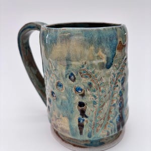 以下が含まれることがあります： ユニークなデザインの手作りセラミックマグ。マグカップは、テクスチャード加工された植物柄と濃い青色のアクセントが施された青とタンの釉薬が特徴です。ハンドルは湾曲しており、マグカップは幅広のベースを備えています。