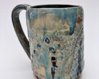 Könnte beinhalten: Eine handgefertigte Keramiktasse mit einem einzigartigen Design. Die Tasse hat eine blaue und beige Glasur mit strukturierten botanischen Mustern und dunkelblauen Akzenten. Der Griff ist gebogen und die Tasse hat einen breiten Boden.
