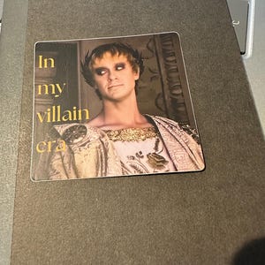 Puede incluir: Una pegatina con un retrato de una persona con vestimenta romana y el texto "In my villain era". La persona lleva una corona de laurel y una prenda con adornos dorados. La pegatina está sobre un cuaderno gris oscuro.