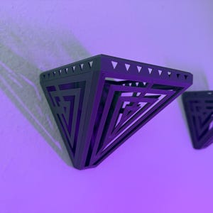 Könnte beinhalten: Zwei dunkelgraue, geometrische Wandregale in Dreiecksform. Die Regale haben ein geschichtetes Design mit ausgeschnittenen Mustern und sind an einer hellvioletten Wand befestigt. Die Regale sind leer.