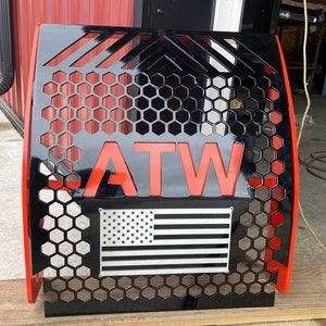 May include: Black metal panel with orange trim and the letters "ATW" in orange. The panel features a honeycomb pattern and a silver and black American flag design. The panel is likely for a vehicle.