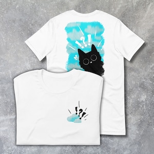 Puede incluir: Camisetas blancas con un diseño de gato. Una camiseta presenta un gato negro con signos de interrogación sobre un fondo azul. La otra tiene un pequeño diseño de nube y signo de interrogación.