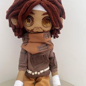 Puede incluir: Muñeca de tela hecha a mano con cabello de hilo marrón, ojos de botón y una cara cosida. La muñeca lleva una chaqueta marrón, pantalones color canela y un cinturón blanco. Tela blanca envuelve las muñecas de la muñeca. Un artículo único, hecho a mano.