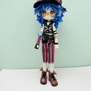 Puede incluir: Muñeca hecha a mano con pelo azul de hilo, que lleva un sombrero negro con gafas, un chaleco, pantalones a rayas y botas marrones. La muñeca tiene una cara detallada y posa sobre un fondo verde claro.